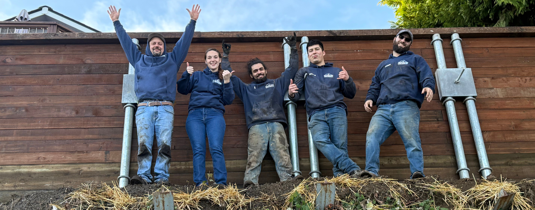 The Rescuers Down Under team (four men and a woman in navy blue hoodies and dirty jeans) standing in front of a large wooden retaining wall